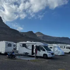 Un grupo de autocaravanas low cost aparcadas en un terreno pedregoso, con montañas y cielo azul de fondo.