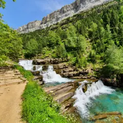 Sendero de piedra junto a las cascadas de las **gradas de soaso**, con vegetación exuberante y un imponente macizo rocoso al fondo.