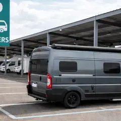 Estacionamiento para campers bajo techo. Un letrero verde indica "CAMPERS". Varios vehículos recreativos, incluyendo una furgoneta gris en primer plano, están aparcados.