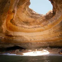 Interior de una cueva marina con un agujero en el techo, donde está Benagil. Gente en kayaks y en la playa disfrutan del sol.