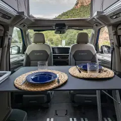 Interior de VW California listo para cenar. Mesa puesta con platos y vasos, asientos delanteros y vistas al exterior.