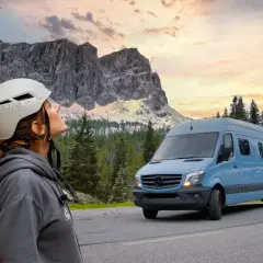 Mujer con casco mira las montañas. A su lado, un **autobús camperizado** azul espera para la aventura.