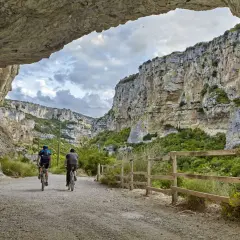 Dos ciclistas exploran la impresionante **foz de Lumbier**, un cañón rocoso con vegetación exuberante y un cielo nublado.