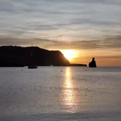 Un velero navega en la serena bahía mientras el sol se pone en Benirràs, tiñendo el cielo de tonos dorados y naranjas.