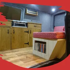 Interior de caravana con cocina, cama y estanterías. El suelo de madera renovado da un aspecto acogedor.