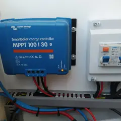 Controlador de carga solar Victron MPPT 100|30, con luces indicadoras de estado y cables conectados. A su derecha, un cuadro eléctrico con interruptores.