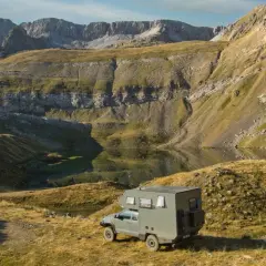 Una camper lista para la aventura en la Transpirenaica, aparcada junto a un lago de montaña con imponentes picos rocosos al fondo.