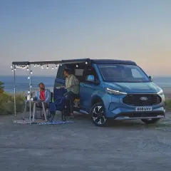 Pareja disfrutando en una playa con una camper hibrida enchufable azul, con toldo y luces.