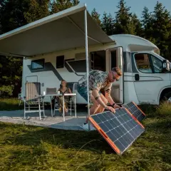 Hombre instala paneles solares, quizás en serie o paralelo, junto a una autocaravana. Mujer trabaja en portátil bajo toldo.