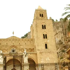 La imponente catedral de Cefalú se alza majestuosa contra un acantilado rocoso, con una palmera que añade un toque tropical a la escena.