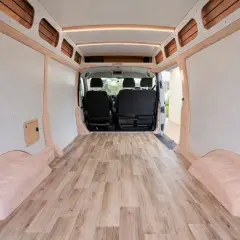Interior de furgoneta camper con suelo de madera. Las paredes están revestidas y hay asientos en la parte delantera.