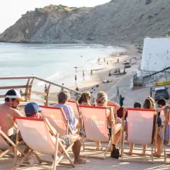 Gente relajándose en tumbonas con vistas a la playa de Burgau. El mar azul y la costa rocosa crean un paisaje idílico.