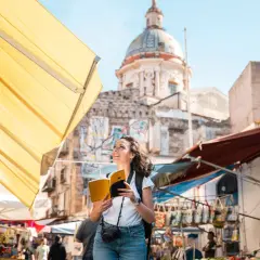 Turista sonriente con libro y cámara explora los vibrantes mercados de Palermo, con un domo histórico al fondo.