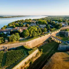 Vista aérea de la ciudadela de Blaye, con sus imponentes murallas de piedra, un puente arqueado y viñedos verdes junto a un río sereno.