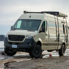 Una van 4x4 color arena, lista para la aventura invernal, con equipo de campamento en el techo y escaleras laterales, aparcada en un paisaje nevado.