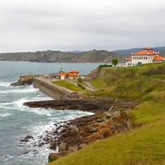 Costa de San Vicente de la Barquera a Comillas con edificios blancos y tejados naranjas, mar agitado y acantilados verdes.