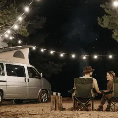 Pareja acampando bajo luces de feria en la costa atlántica francesa. Una furgoneta camper y sillas plegables crean un ambiente acogedor.