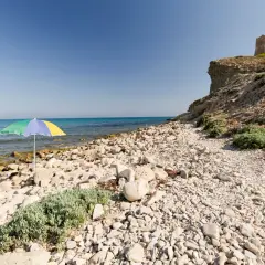 Sombrilla de colores en la playa de las covaticas, con mar turquesa y torre antigua en el acantilado.