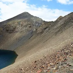 Senderistas ascienden por un sendero pedregoso junto a un lago azul, con la imponente silueta de los ibones de Bachimaña al fondo.