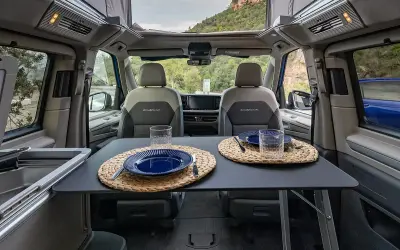 Interior de VW California listo para cenar. Mesa puesta con platos y vasos, asientos delanteros y vistas al exterior.