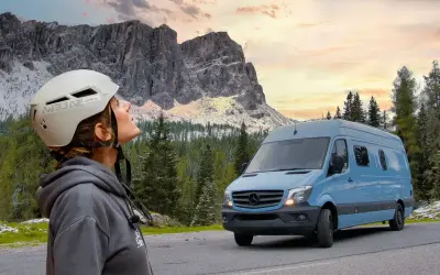 Mujer con casco mira las montañas. A su lado, un **autobús camperizado** azul espera para la aventura.