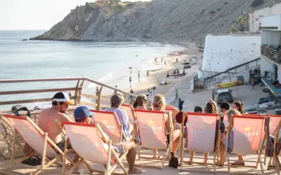 Gente relajándose en tumbonas con vistas a la playa de Burgau. El mar azul y la costa rocosa crean un paisaje idílico.