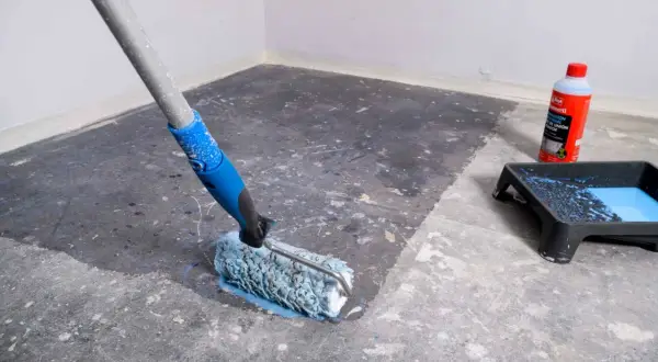 Rodillo aplicando líquido para quitar sintasol del suelo. Bandeja con pintura azul y botella de producto al lado.