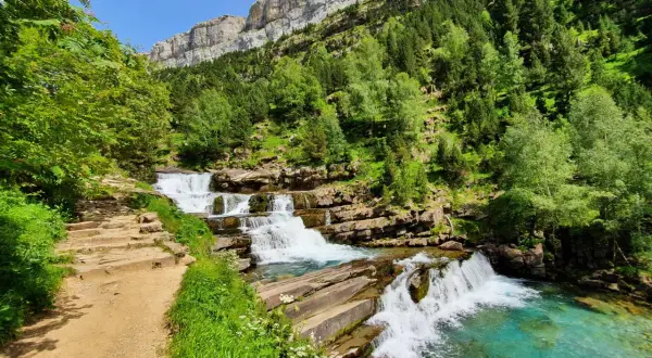 Sendero de piedra junto a las cascadas de las **gradas de soaso**, con vegetación exuberante y un imponente macizo rocoso al fondo.