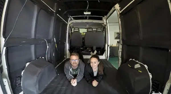 Pareja sonríe tumbada en el suelo de una furgoneta camper en proceso de aislamiento. Merece la pena aislar furgoneta camper para futuras aventuras.
