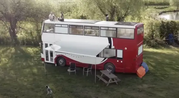 Autobús motorhome de dos pisos con paneles solares en el techo. Personas sentadas en la parte superior, un toldo extendido y muebles de exterior.