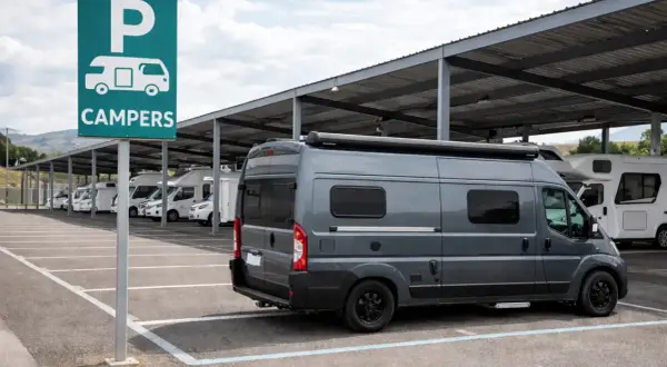 Estacionamiento para campers bajo techo. Un letrero verde indica "CAMPERS". Varios vehículos recreativos, incluyendo una furgoneta gris en primer plano, están aparcados.