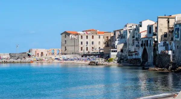 Edificios antiguos y una playa animada en Cefalú, Italia, con el mar azul brillante.