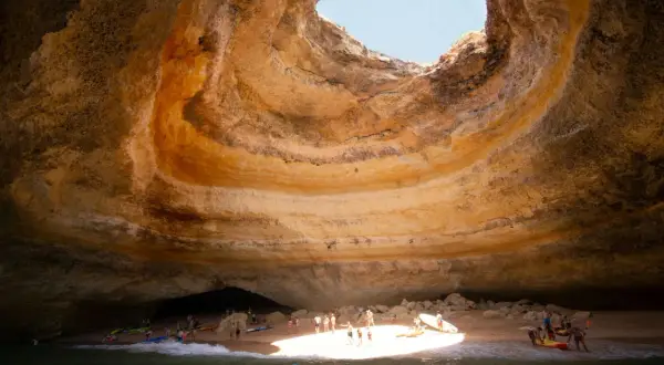 Interior de una cueva marina con un agujero en el techo, donde está Benagil. Gente en kayaks y en la playa disfrutan del sol.