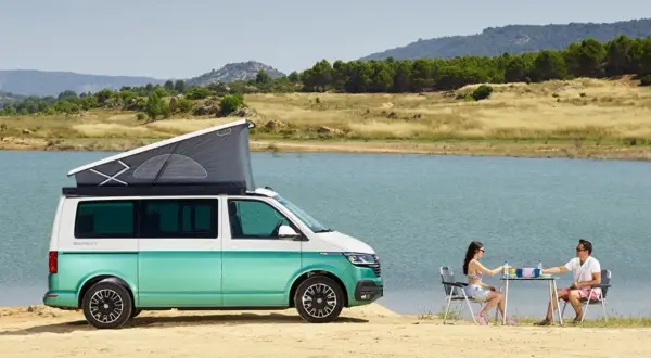Volkswagen California con baño, lista para una escapada junto al lago. Pareja disfruta de un picnic al aire libre.
