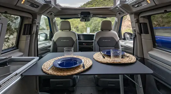 Interior de VW California listo para cenar. Mesa puesta con platos y vasos, asientos delanteros y vistas al exterior.