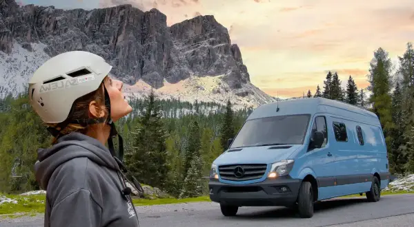 Mujer con casco mira las montañas. A su lado, un **autobús camperizado** azul espera para la aventura.