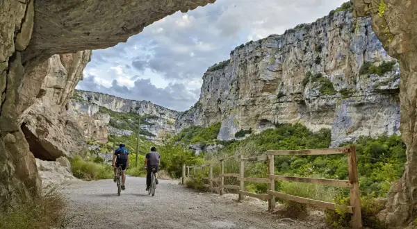 Dos ciclistas exploran la impresionante **foz de Lumbier**, un cañón rocoso con vegetación exuberante y un cielo nublado.