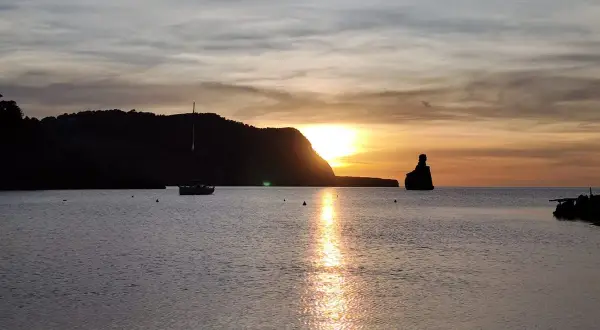 Un velero navega en la serena bahía mientras el sol se pone en Benirràs, tiñendo el cielo de tonos dorados y naranjas.