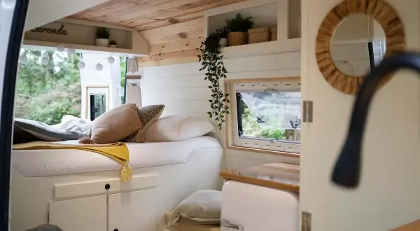 Interior de una camper con cama acogedora, cojines y mantas. Muestra cómo llevar la ropa de forma organizada en este espacio.