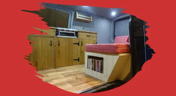 Interior de caravana con cocina, cama y estanterías. El suelo de madera renovado da un aspecto acogedor.