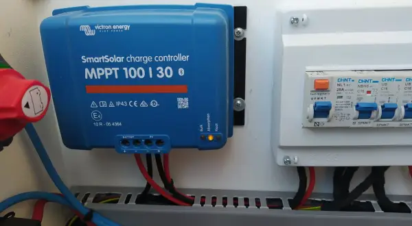 Controlador de carga solar Victron MPPT 100|30, con luces indicadoras de estado y cables conectados. A su derecha, un cuadro eléctrico con interruptores.