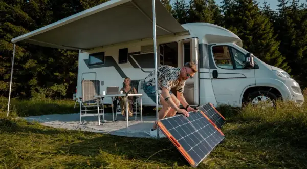 Hombre instala paneles solares, quizás en serie o paralelo, junto a una autocaravana. Mujer trabaja en portátil bajo toldo.
