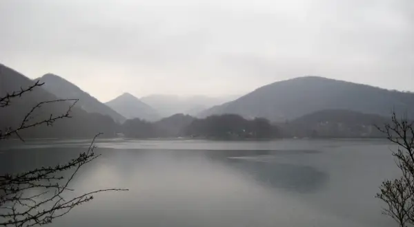 Paisaje brumoso con montañas cubiertas de árboles y el **embalse de Lareo** en primer plano. Ramas desnudas enmarcan la serena vista.