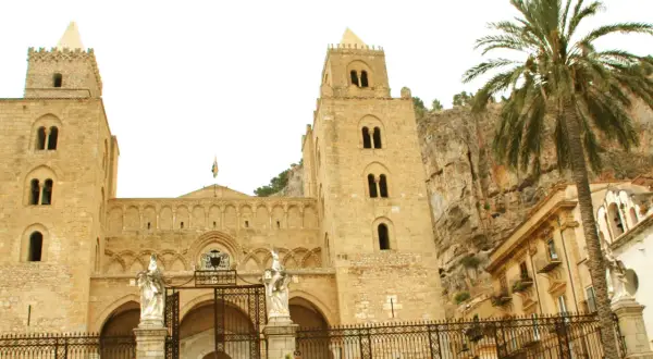 La imponente catedral de Cefalú se alza majestuosa contra un acantilado rocoso, con una palmera que añade un toque tropical a la escena.