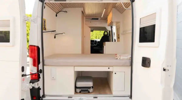 Interior de una camper con cama, almacenamiento y un pequeño equipaje. La altura camper permite aprovechar cada rincón.