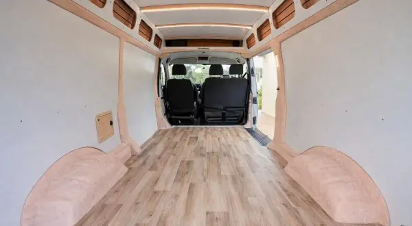Interior de furgoneta camper con suelo de madera. Las paredes están revestidas y hay asientos en la parte delantera.