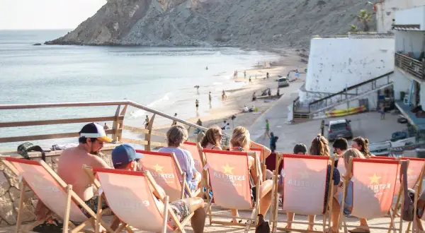 Gente relajándose en tumbonas con vistas a la playa de Burgau. El mar azul y la costa rocosa crean un paisaje idílico.