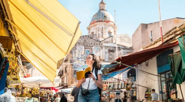 Turista sonriente con libro y cámara explora los vibrantes mercados de Palermo, con un domo histórico al fondo.