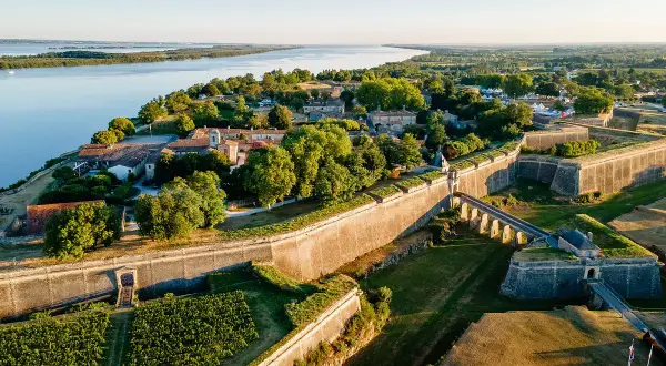 Vista aérea de la ciudadela de Blaye, con sus imponentes murallas de piedra, un puente arqueado y viñedos verdes junto a un río sereno.