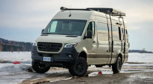 Una van 4x4 color arena, lista para la aventura invernal, con equipo de campamento en el techo y escaleras laterales, aparcada en un paisaje nevado.